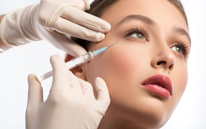 Serine young woman is getting facial botox injection. Beautician hands in gloves holding syringe near her face